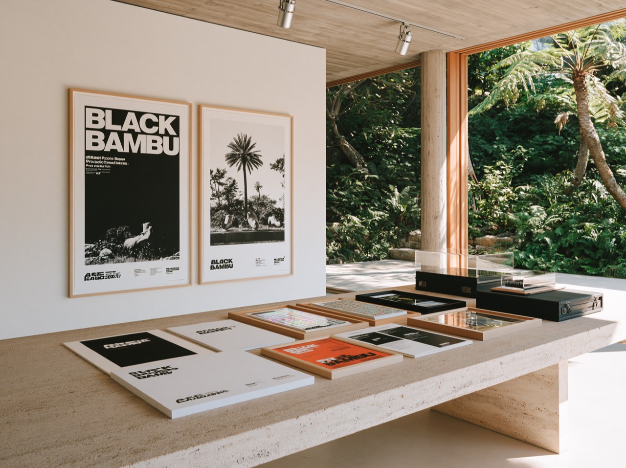 Black Bambu posters, material samples, and archival cases displayed on a stone gallery table beside a tropical garden